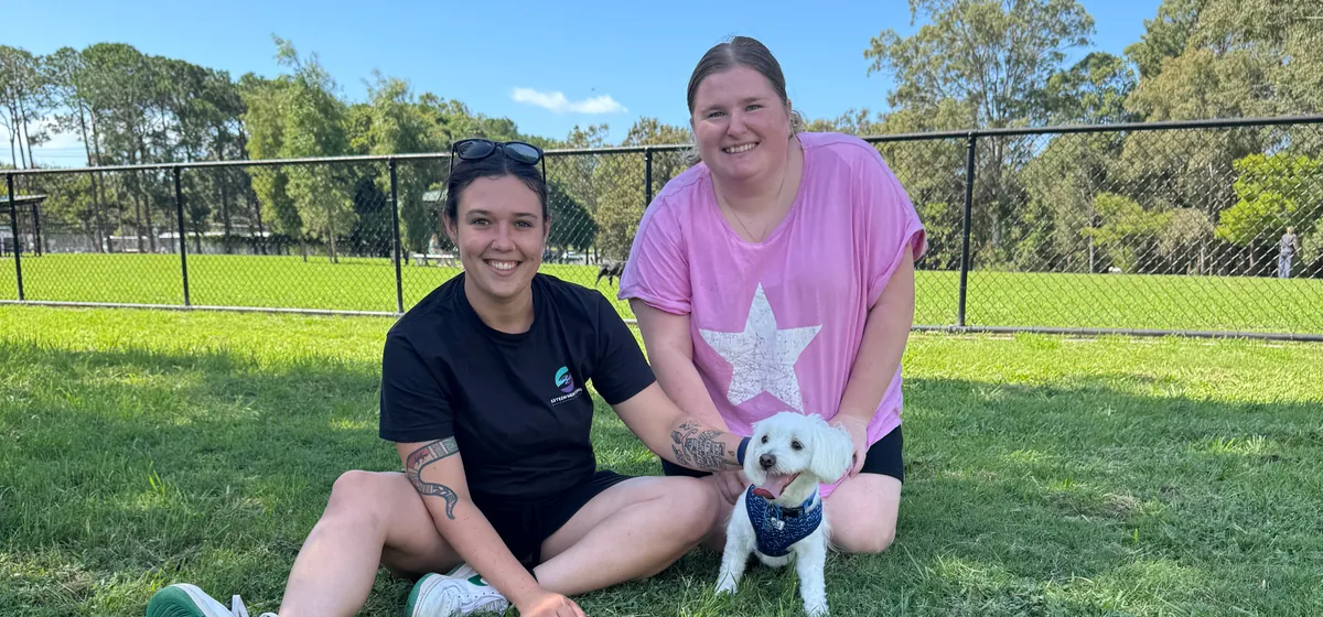 Two Esteem Mentoring team members smiling on the grass at a local park with a small white dog